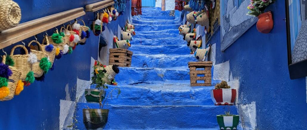 Scalinata azzurra in un vicolo stretto della medina di Chefchaouen, Marocco, decorata con ceste di paglia con pompon, quadri, tende a frange, vasi e una porta blu in cima.