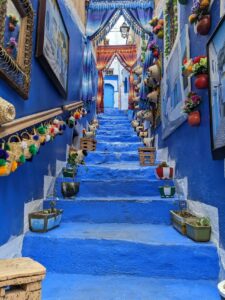 Scalinata azzurra in un vicolo stretto della medina di Chefchaouen, Marocco, decorata con ceste di paglia con pompon, quadri, tende a frange, vasi e una porta blu in cima.
