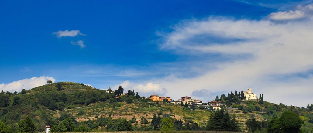 Campi e colline terrazzate di Montevecchia in Brianza, con case e una chiesa sul crinale sotto il cielo azzurro