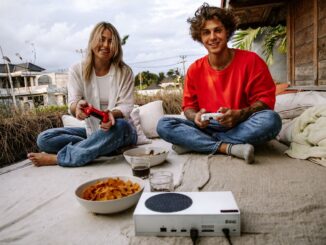 Due persone giocano ai videogiochi all'aperto davanti a uno schermo, con un paesaggio rurale sullo sfondo