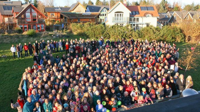 Foto di gruppo della comunità di Findhorn in Scozia, sul prato davanti alle case con grandi finestre e pannelli solari.